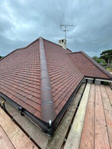 View from scaffolding of a ridged new roof fitted with flat brick coloured tiles and a ridge with clay tiles