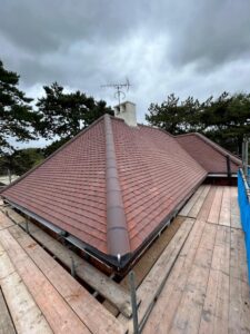 View from scaffolding of a ridged new roof fitted with flat brick coloured tiles and a ridge with clay tiles