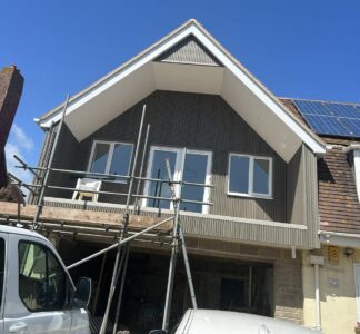 View of a work in progress from ground level of a two story extension with pitched roof, windows and a balcony with scaffolding in place.