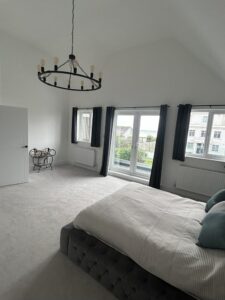 A large bedroom with white walls, grey carpet and double bed, with overhanging chandalier and french doors leading to a balcony.